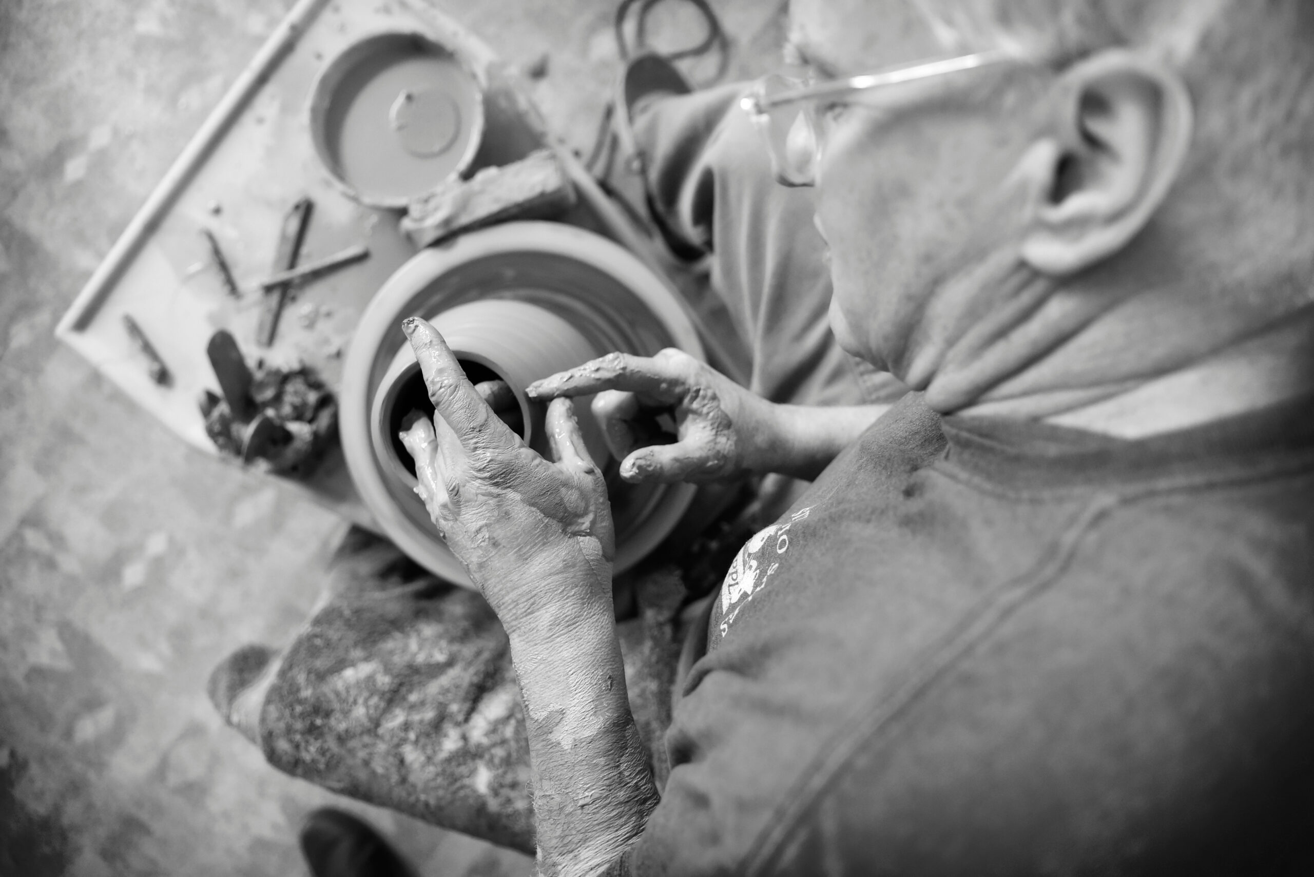 Black and white photo over the shoulder of Mike Carrol working with clay on a pottery wheel.